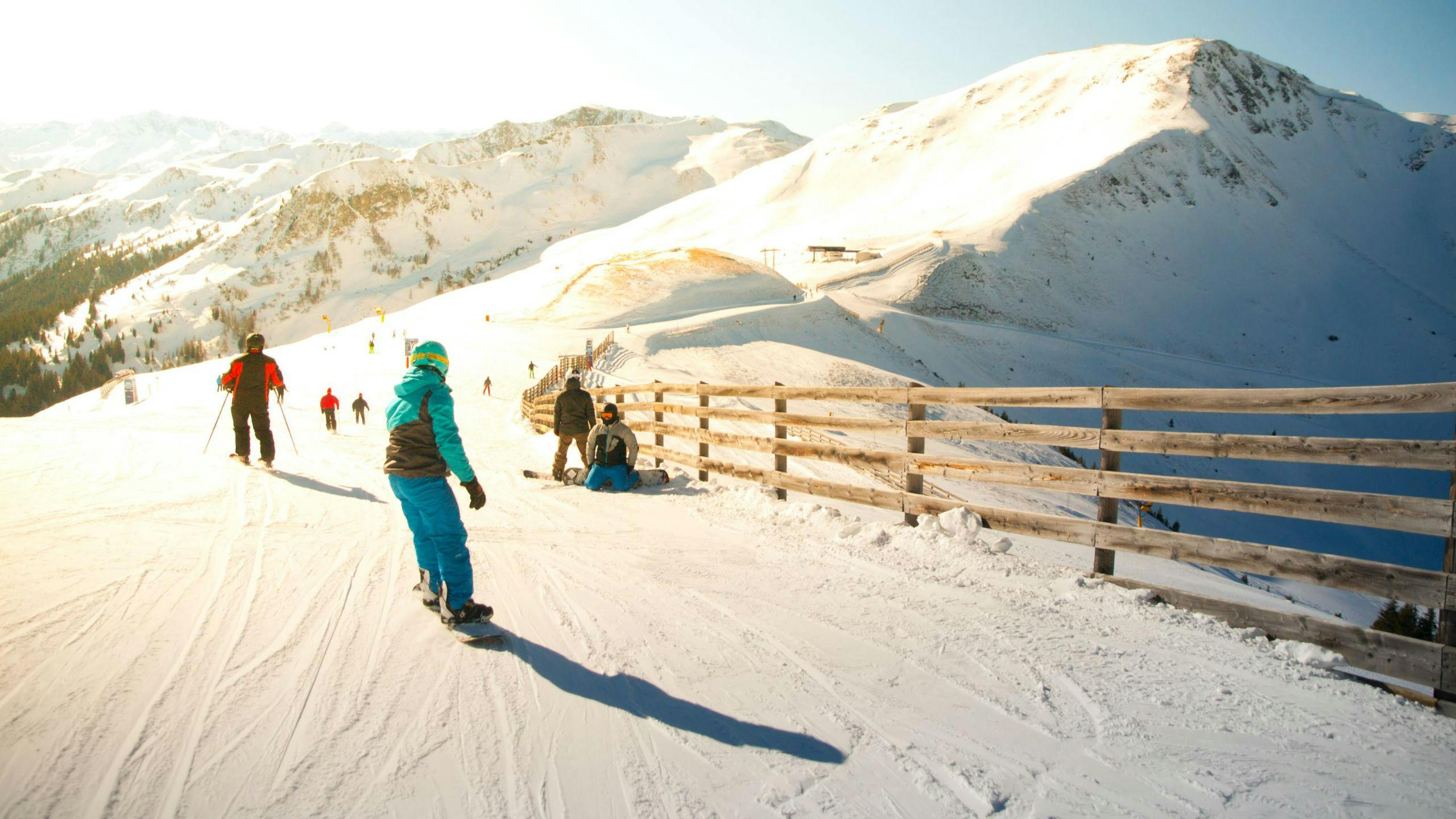A couple of skiers and snowboarders are riding down a sunlit skislope in the ski resort of Saalbach, where local ski schools offer ski lessons for people who want to learn to ski.